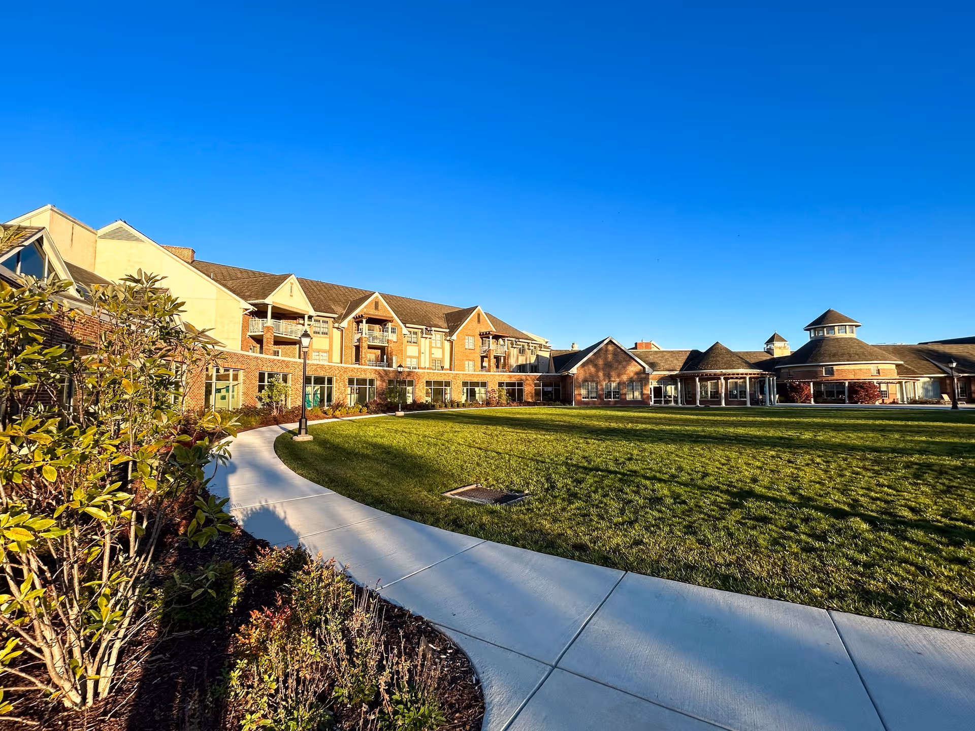 Wide exterior view of Cross Keys Village senior living facility showing a large green lawn, curved concrete walkway, landscaped bushes, and a multi-story brick building under a clear blue sky.