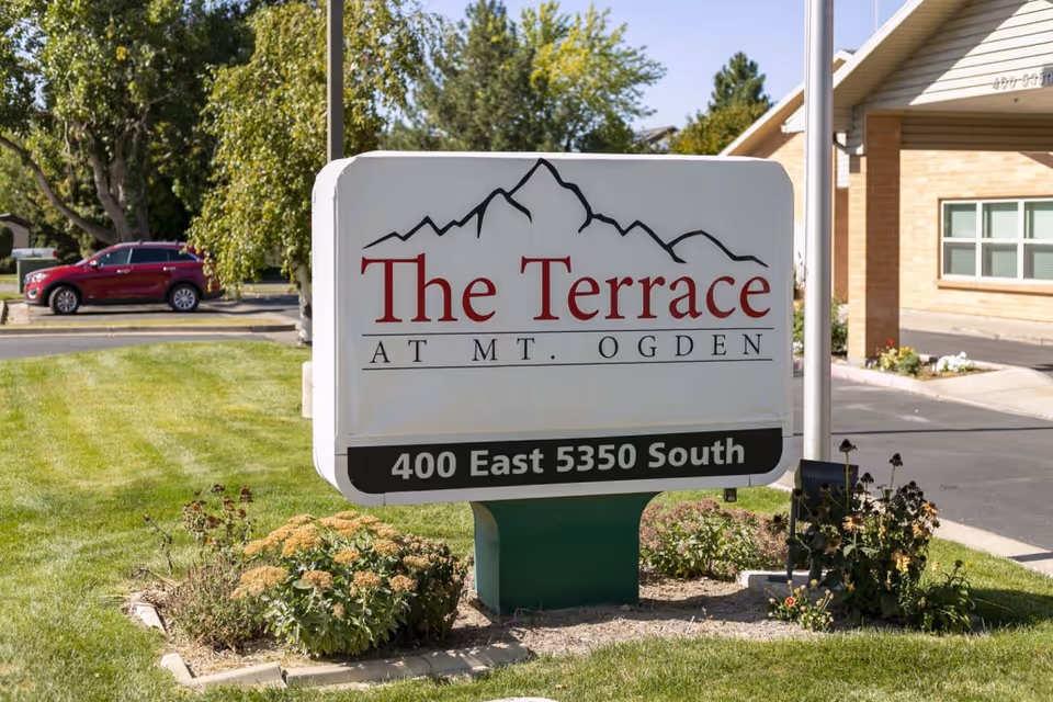 Outdoor view of a sign for The Terrace at Mt. Ogden, located at 400 East 5350 South, surrounded by grass, plants, and trees with a building and a red car in the background.