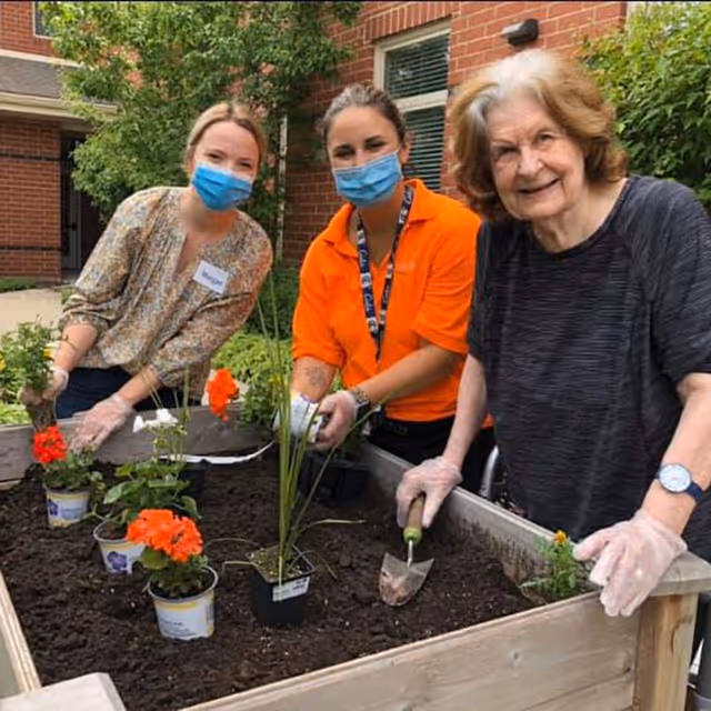 Three women, two wearing face masks and gloves, are gardening together in a raised garden bed outside a brick building. They are planting flowers and smiling at the camera.