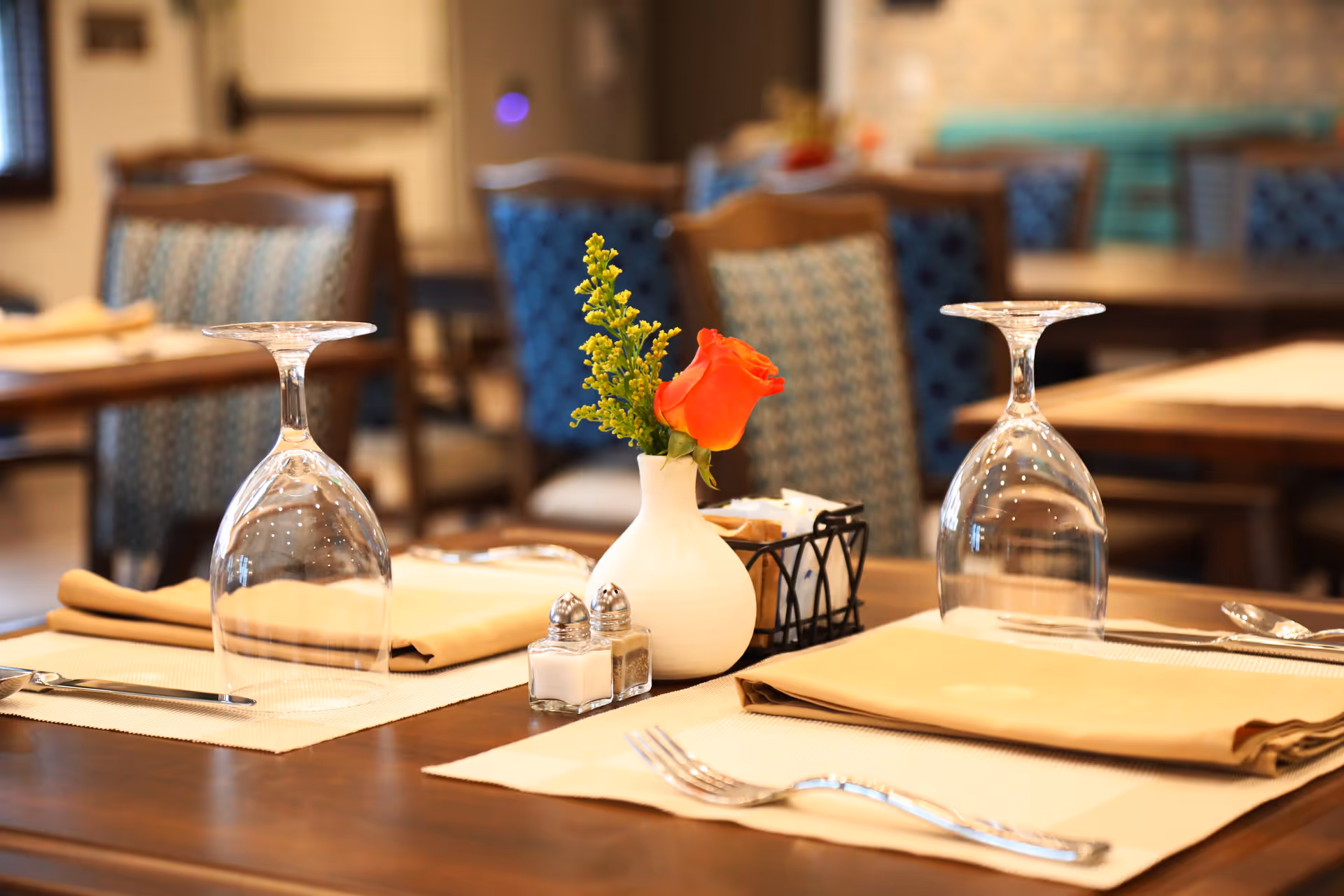 Close-up of a dining table set with upside-down wine glasses, beige napkins, silverware, a small white vase with an orange rose and greenery, and salt and pepper shakers in a dining room with wooden chairs upholstered in patterned fabric.