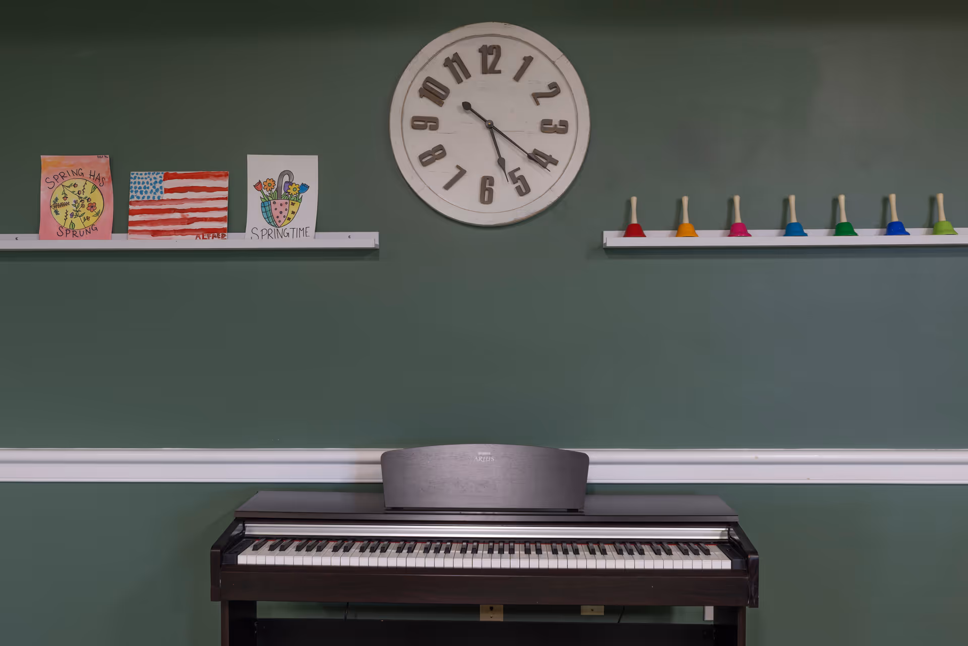 A digital piano against a green wall with a large round clock above it. On the wall, there are two white shelves: one holding three colorful drawings including an American flag and spring-themed artwork, and the other holding a row of colorful handbells.