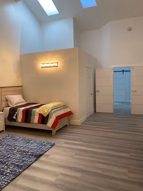 A spacious bedroom with a single bed covered with a striped blanket in red, black, gray, and white colors, and a yellow throw. The room has light-colored walls, a wooden floor, a blue patterned rug, and two skylights on the ceiling. There is a modern wall light above the bed and a doorway leading to another room with white double doors.