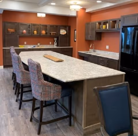 Communal kitchen with a large central island, upholstered bar stools, wood cabinetry and a refrigerator against an orange wall.