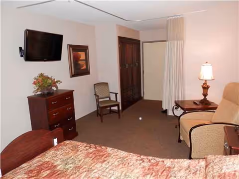Bedroom in a senior living facility with bed, dresser, TV, seating, and closet.