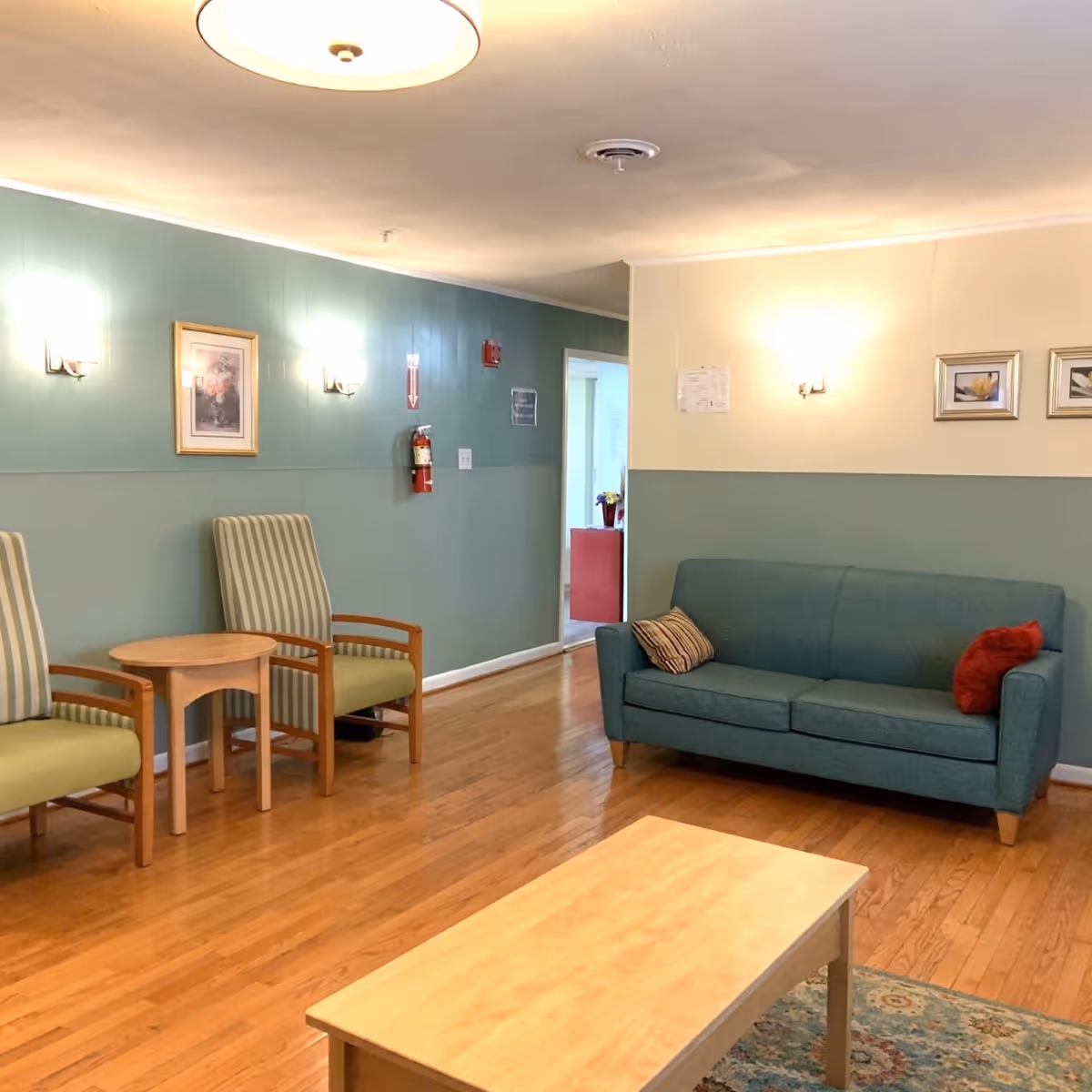 A cozy living room area with wooden flooring, a teal sofa with two cushions, two striped armchairs with wooden arms, a small round wooden table between the chairs, a rectangular wooden coffee table, and framed artwork on the walls painted in two tones of green and beige. The room is softly lit by wall sconces and a ceiling light.