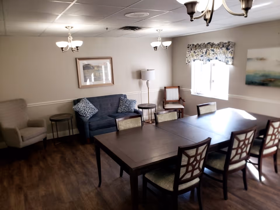 A well-lit interior room with a large wooden dining table surrounded by six chairs. The room features a blue loveseat with two patterned pillows, a beige armchair, a wooden chair, two side tables, a floor lamp, and wall art. There are two ceiling light fixtures and a window with floral curtains letting in natural light.
