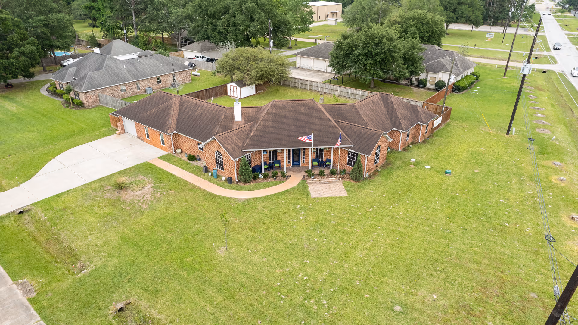 Aerial view of a single-story brick building with a brown roof, surrounded by a large grassy area. The building has a curved driveway and a small front porch with two American flags. There are trees and other buildings in the background, along with a road running alongside the property.