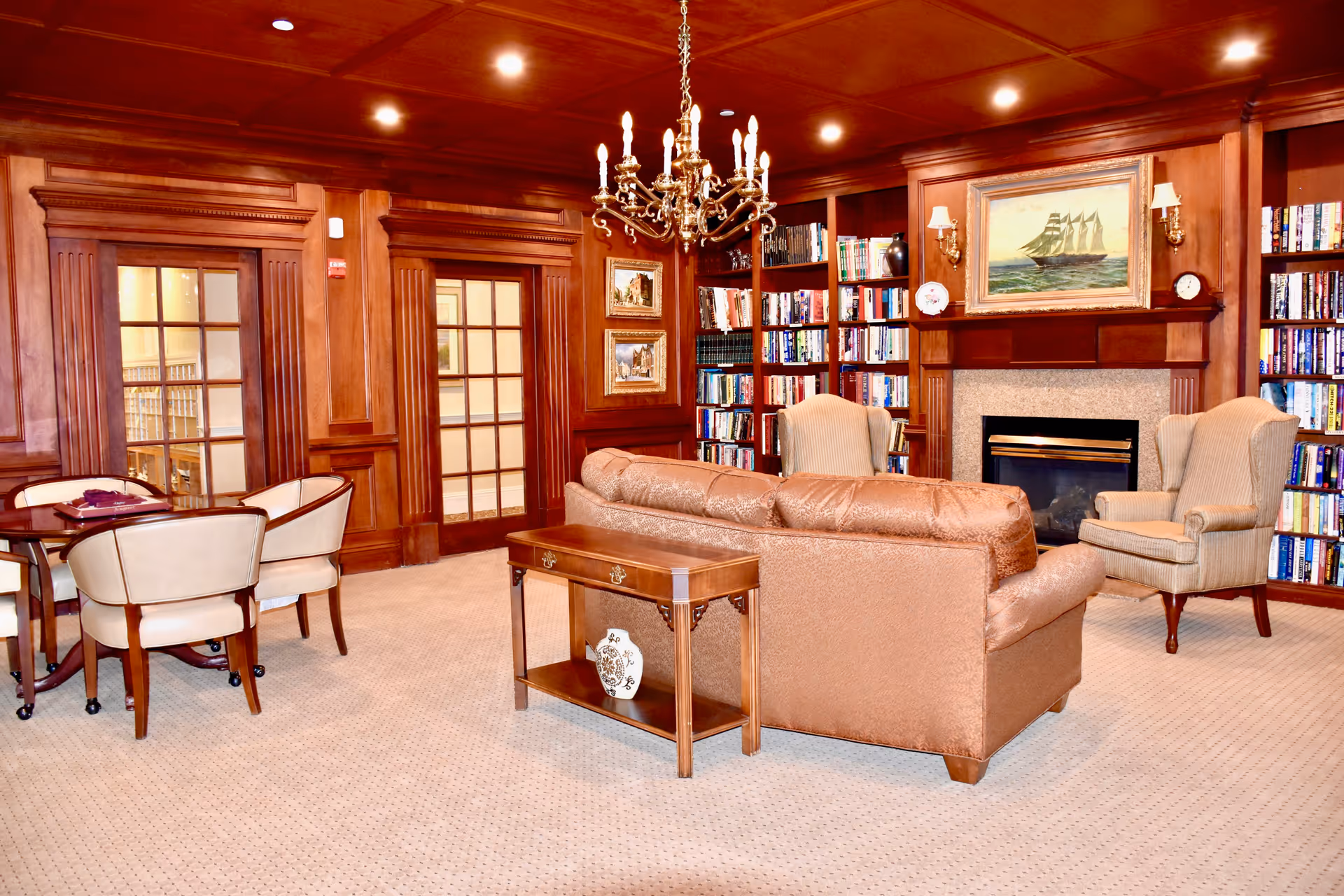 A cozy living room with wood-paneled walls and ceiling, featuring a brown leather sofa, two beige armchairs, a wooden coffee table, and built-in bookshelves filled with books. There is a fireplace with a painting of a sailing ship above it, and a chandelier hanging from the ceiling. Two glass-paneled wooden doors are visible on the left side of the room.