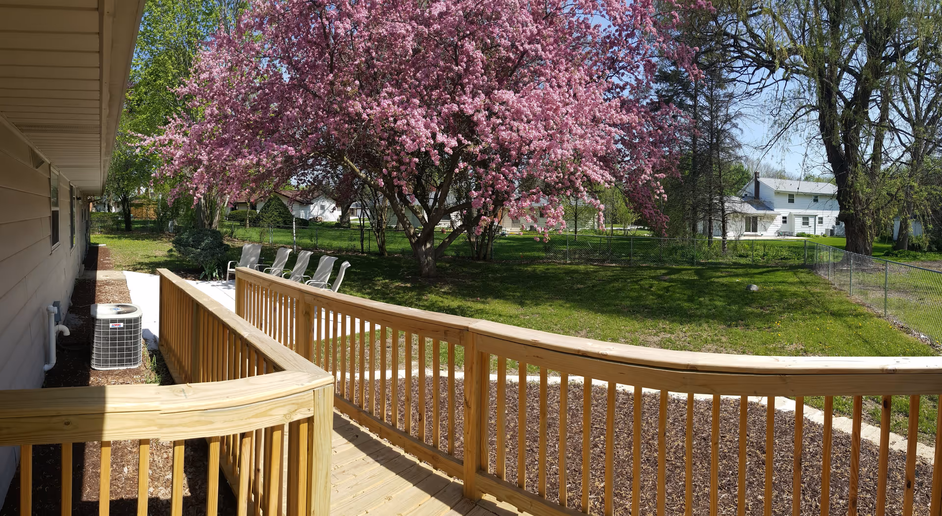 Outdoor view of a senior living facility showing a wooden ramp with railings leading to a patio area with several white chairs. A large tree with pink blossoms is in the grassy yard, surrounded by other trees and a chain-link fence. Nearby houses are visible in the background under a clear blue sky.