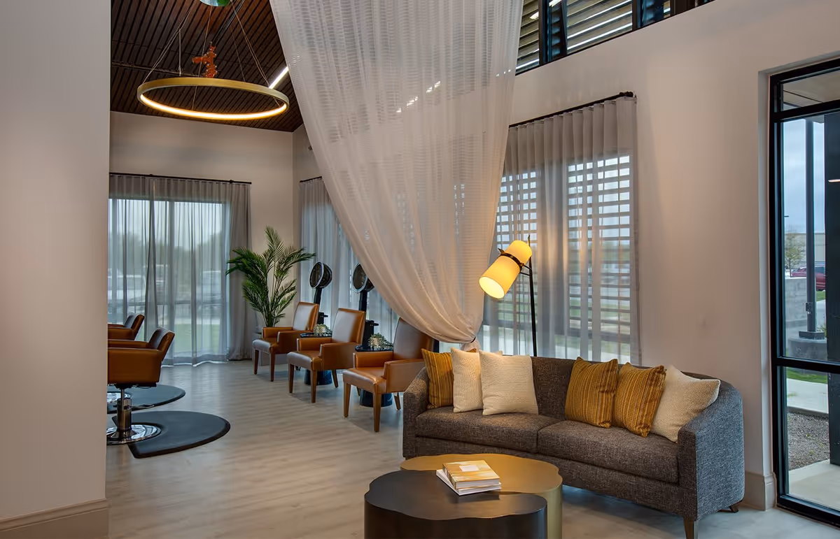 A modern, cozy lounge area with a gray sofa adorned with white and mustard yellow pillows, a floor lamp, and two nested coffee tables with books on top. The space features large windows with sheer curtains, several brown leather chairs, and a tall green plant. The ceiling has a wooden finish with a circular hanging light fixture.