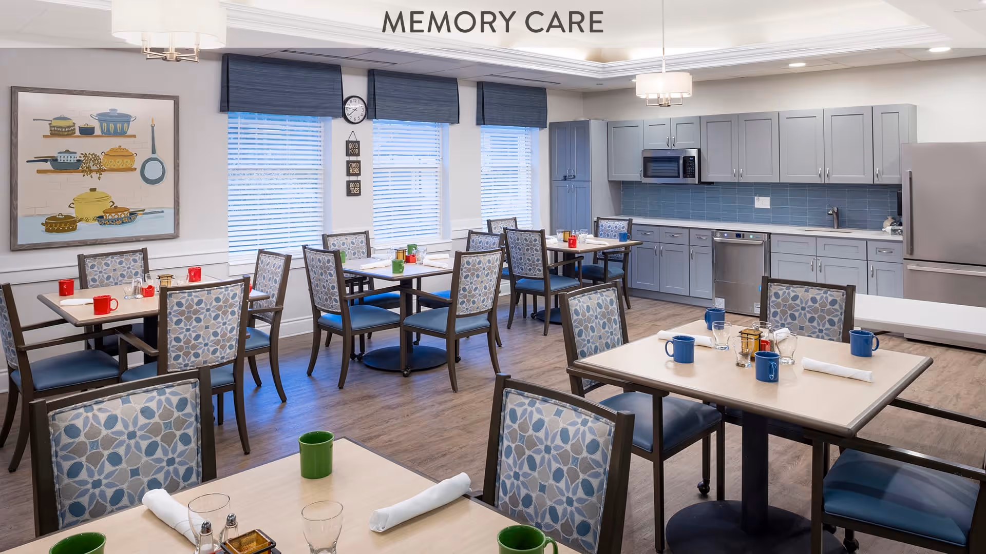 A bright and clean memory care dining room with several tables and chairs featuring patterned upholstery. The room includes a kitchen area with gray cabinets, a microwave, dishwasher, and refrigerator. The tables are set with colorful mugs, glasses, and napkins. Large windows with blue valances allow natural light to fill the space. A framed artwork of kitchen pots and pans hangs on the wall.