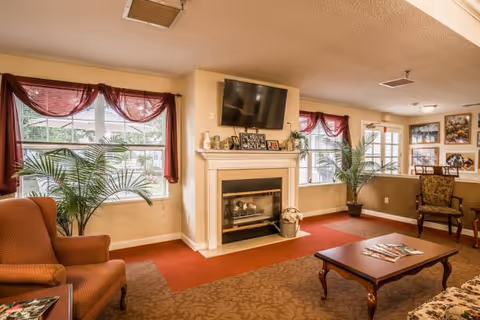 A cozy living room area with a fireplace in the center, a flat-screen TV mounted above it, two large windows with red curtains, potted plants on either side of the fireplace, an armchair on the left, a wooden coffee table with magazines, and a chair with floral upholstery on the right.