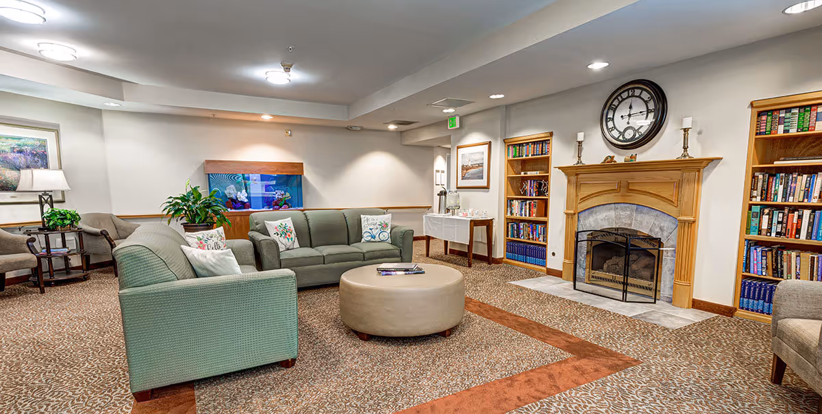 A cozy living room area in a senior living facility featuring two green sofas with decorative pillows, a round beige ottoman, a fireplace with a wooden mantel, two bookshelves filled with books, a large wall clock above the fireplace, a fish tank, and a small table with refreshments in the background.