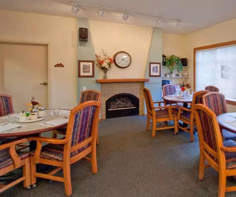 A cozy dining room with round tables and wooden chairs upholstered in striped fabric. The room features a fireplace with a clock and floral arrangement above it, framed pictures on the walls, and a large window with blinds letting in natural light.