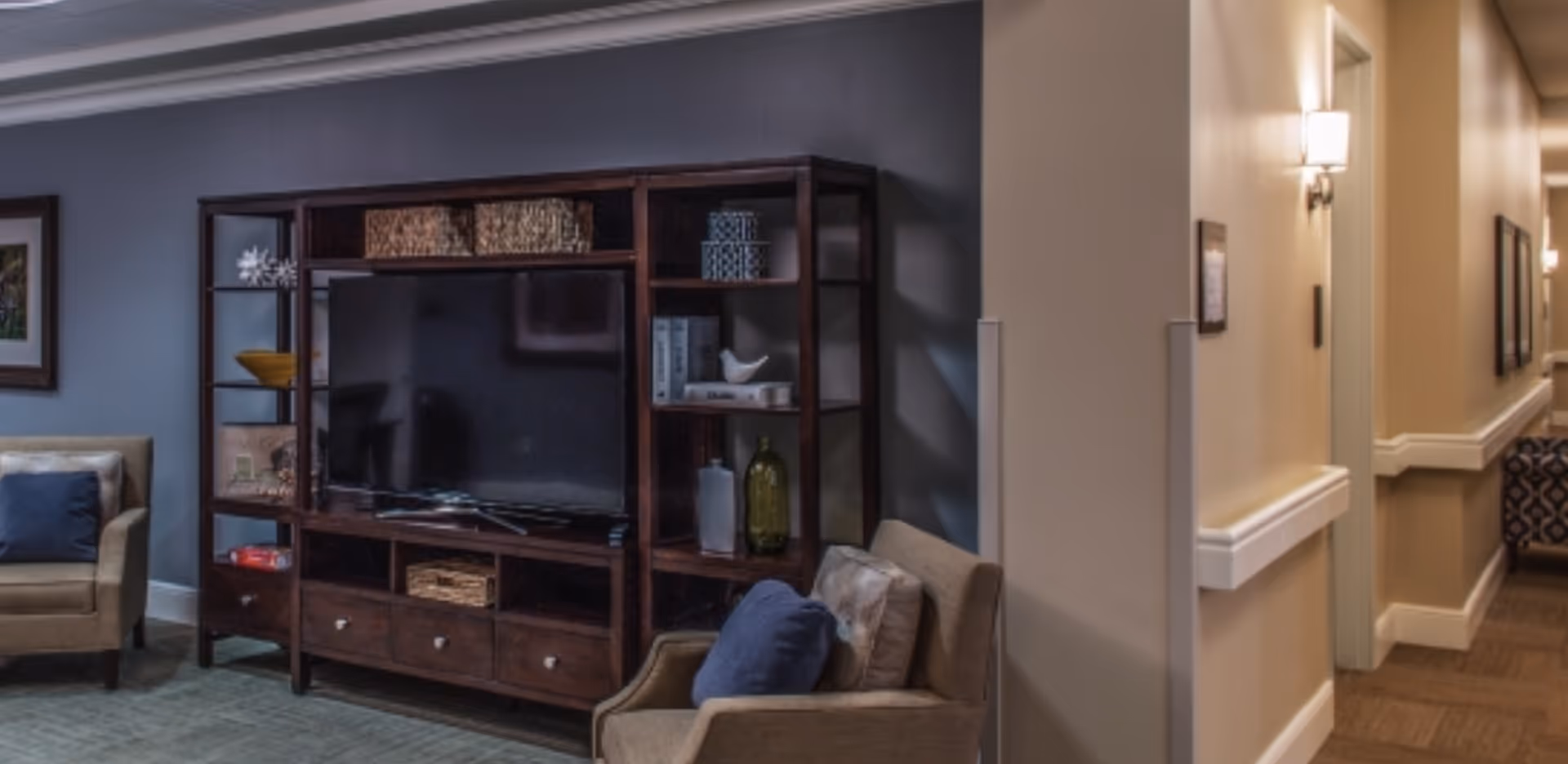 A cozy living room area with a large dark wood entertainment center holding a flat-screen TV and decorative items. There are two beige armchairs with blue and beige cushions positioned near the entertainment center. The room has a gray wall and a carpeted floor. To the right, there is a hallway with beige walls, white trim, and wall-mounted lights.