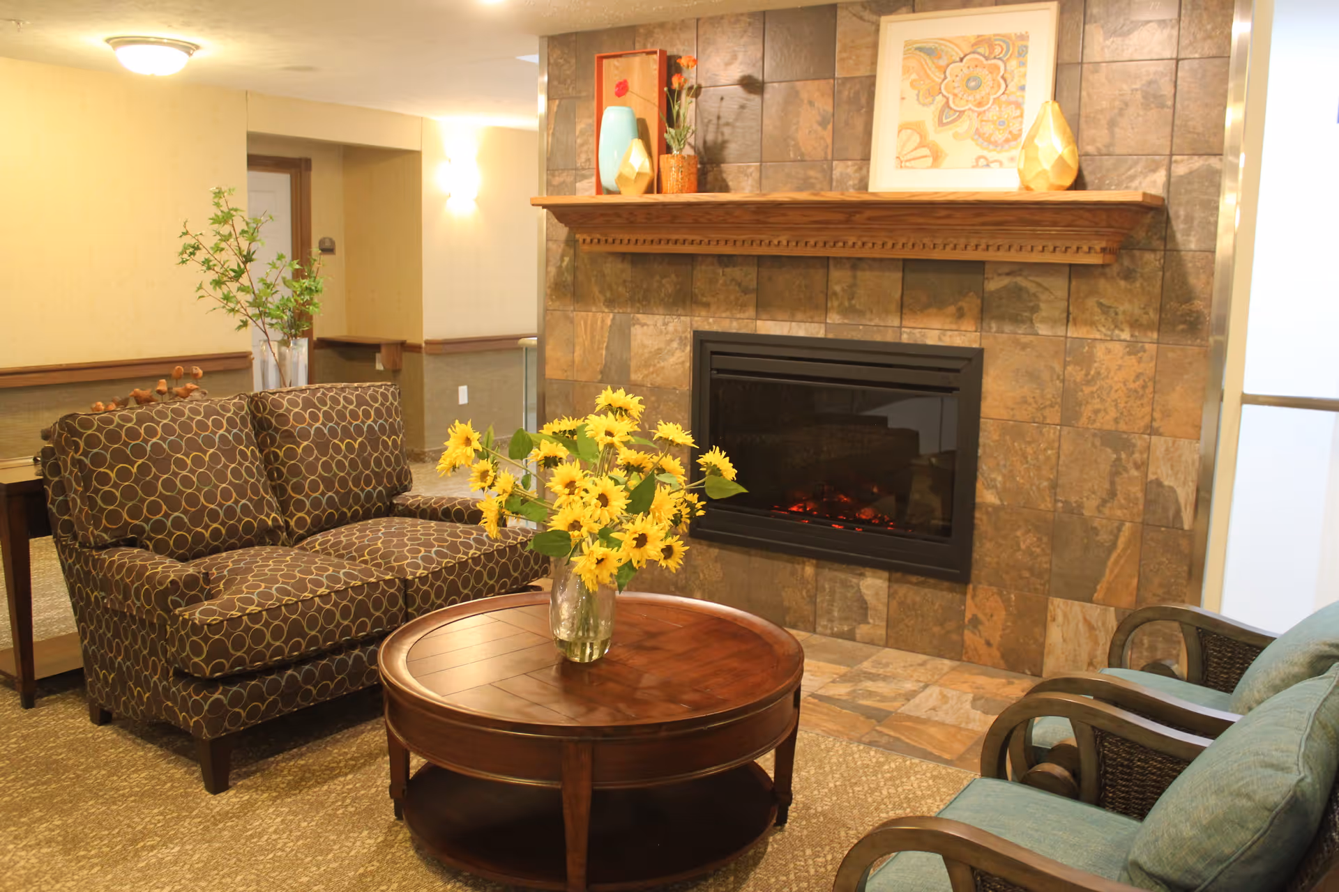 A cozy living room area with a patterned loveseat, two blue cushioned chairs, and a round wooden coffee table with a vase of yellow flowers. The background features a stone-tiled fireplace with a wooden mantel decorated with vases and framed artwork.