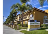 Front view of a three-story Mediterranean-style senior apartment building with palm trees, balconies, and a Huntington Villas sign on the lawn.