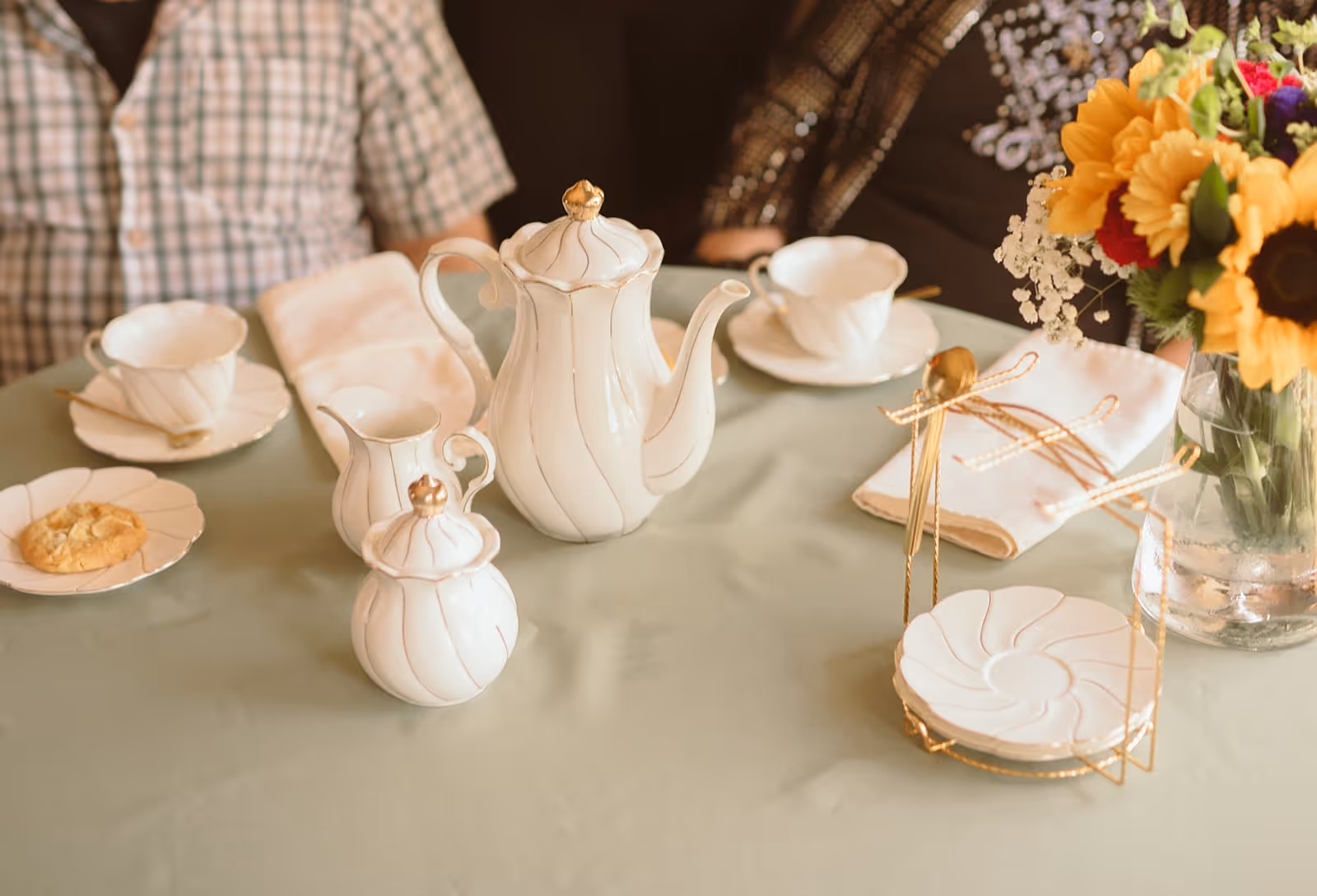 A table set for tea with a white porcelain tea set including a teapot, creamer, sugar bowl, cups, and saucers. There is a cookie on one saucer, folded white napkins with gold spoons, and a vase with a colorful bouquet of flowers including sunflowers. Two people are partially visible sitting at the table.
