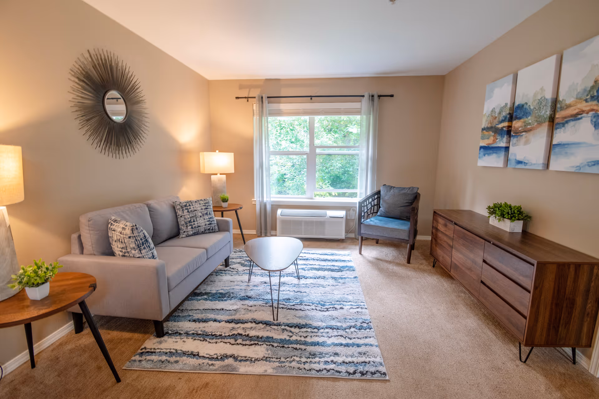 Sunlit living room with a gray sofa, armchair, coffee table on a patterned rug, wooden side table and sideboard, and a window overlooking greenery.