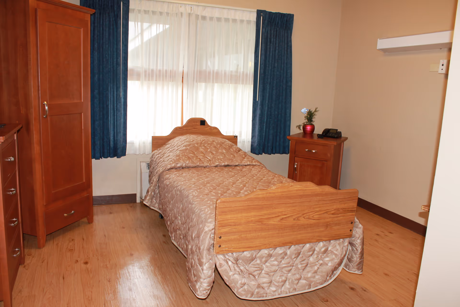 A single bed with a wooden headboard and footboard covered with a beige quilted bedspread in a room with wooden flooring. Next to the bed is a wooden nightstand with a red vase holding a small plant and a telephone. There is a wooden wardrobe and dresser on the left side of the room. A window with sheer white curtains and dark blue drapes is behind the bed.