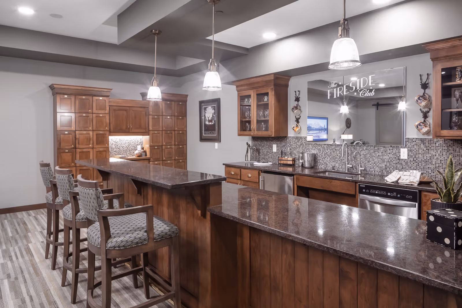 A modern kitchen area with dark granite countertops and wooden cabinetry. There are four cushioned bar stools along a long counter, pendant lights hanging from the ceiling, a mosaic tile backsplash, and a large mirror with the words 'The Fireside Club' on it. The space is well-lit and has a cozy, inviting atmosphere.