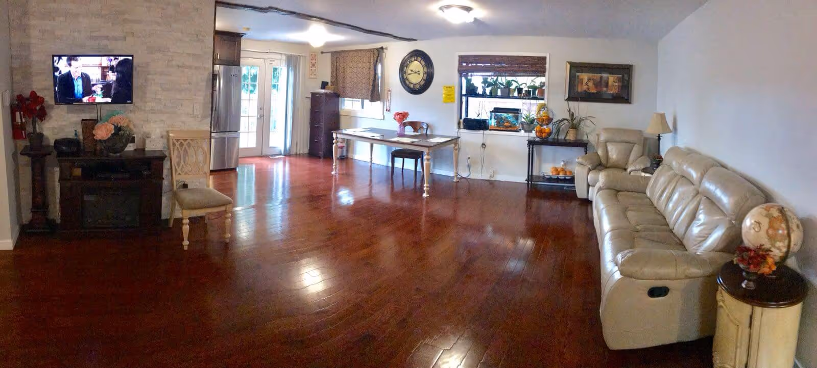 Open living room with polished hardwood floors, a beige leather sofa on the right, a wall-mounted TV on the left, and a table and kitchenette by French doors in the background.