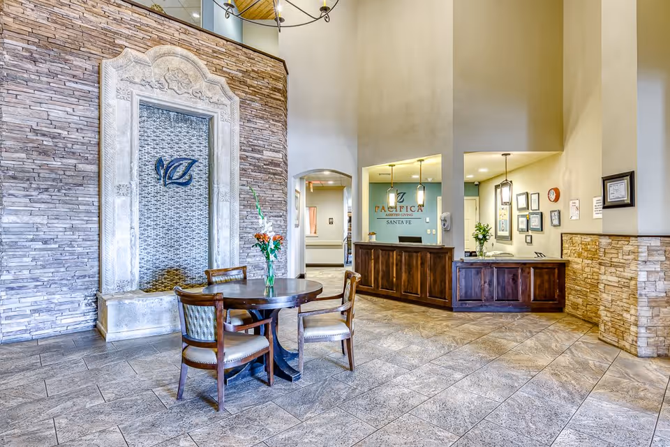Bright assisted-living reception lobby with a round table and chairs, a decorative stone fountain wall, and a wooden front desk bearing the 'Pacifica Assisted Living Santa Fe' logo.