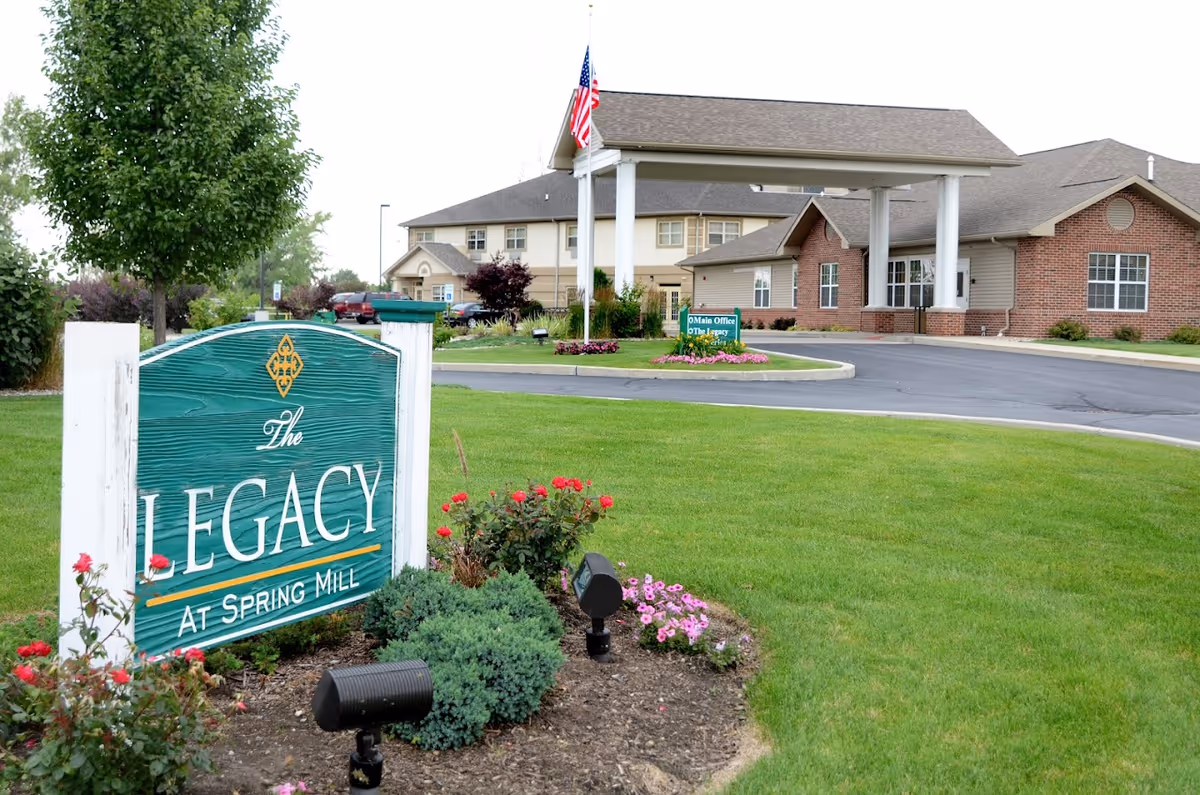 Exterior view of The Legacy at Spring Mill senior living facility showing a green sign with the facility name, a well-maintained lawn with flowers and shrubs, an American flag on a flagpole, and a building with a covered entrance and multiple windows.