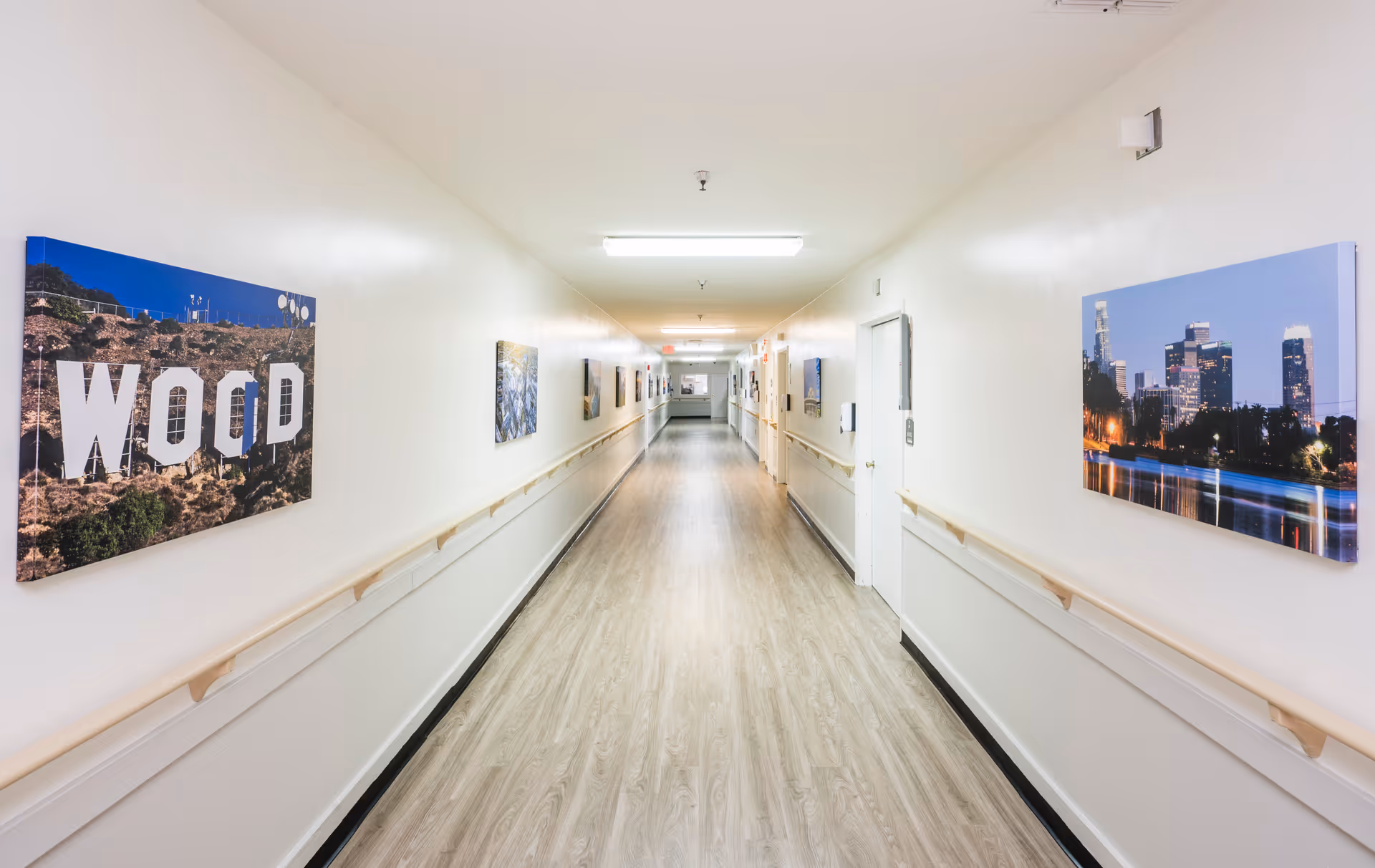 A long, well-lit hallway with light-colored walls and wood-patterned flooring. Handrails run along both sides of the corridor. Several framed pictures, including one of the Hollywood sign and a city skyline at night, are hung on the walls.