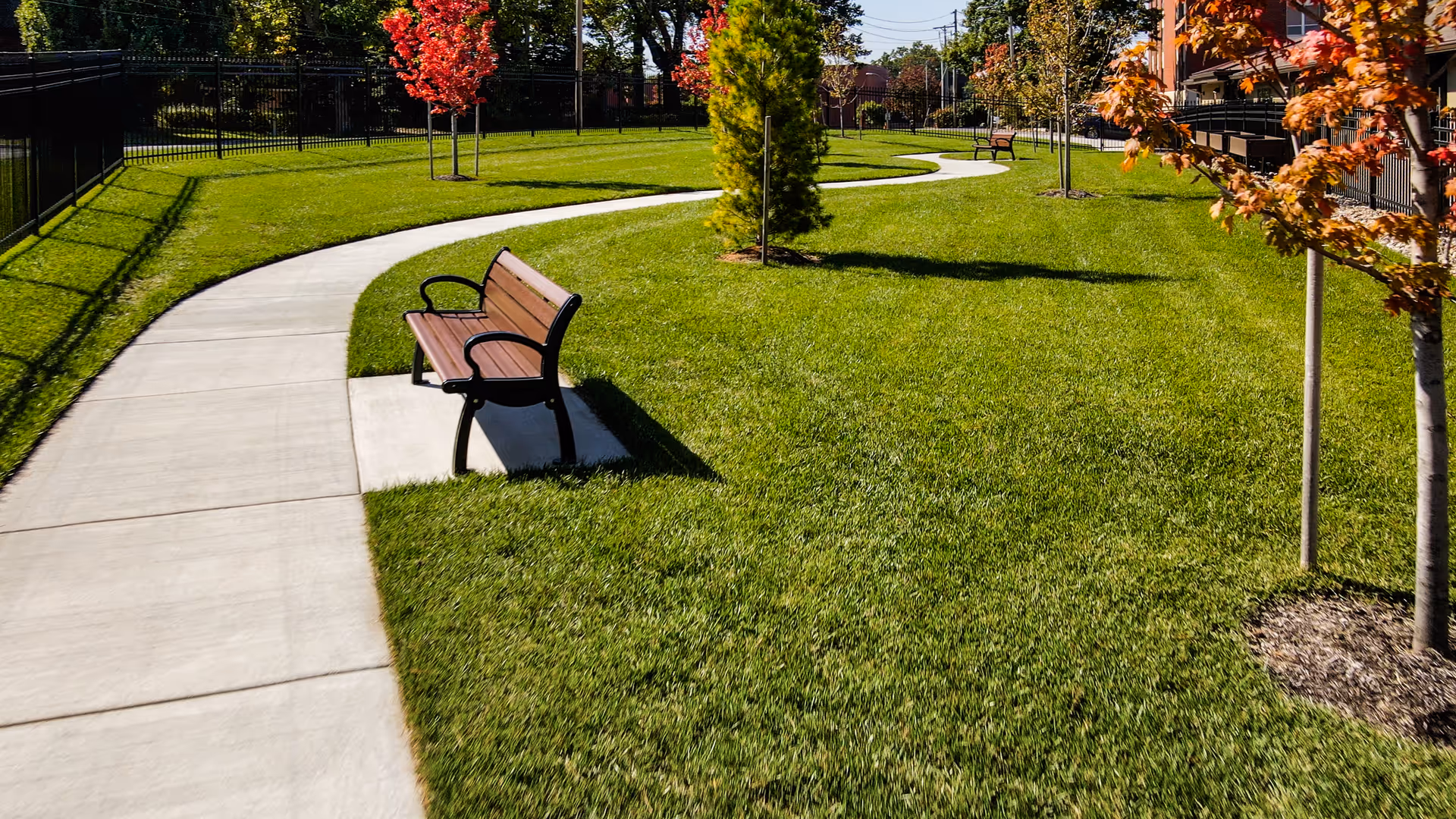 A winding concrete pathway through a well-maintained grassy area with several wooden benches and young trees with autumn-colored leaves. A black metal fence runs along the left side, and a building with balconies is visible on the right side.