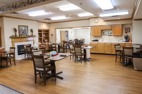 A spacious dining area in a senior living facility with multiple wooden tables and chairs arranged neatly. The room features a fireplace with decorative items on the mantel, wooden cabinetry along one wall, and bright overhead lighting. The floor is wood, and the walls are painted a light color with a decorative border near the ceiling.