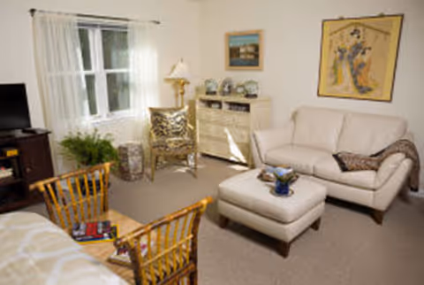 A cozy living room with a white sofa, matching ottoman, two wooden chairs with cushions, a wooden TV stand with a television, a small cabinet with decorative items, a floor lamp, and framed artwork on the walls. A window with sheer white curtains allows natural light into the room.