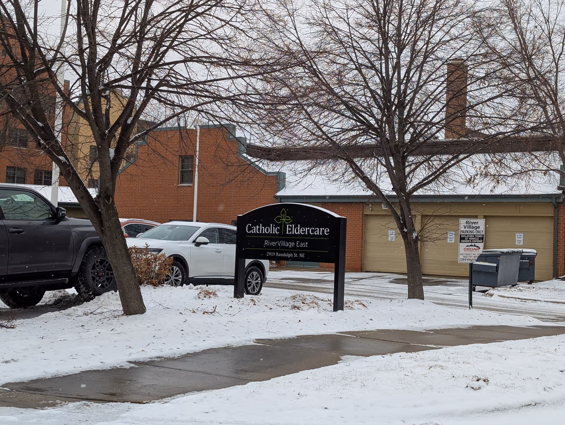 Snow-covered sidewalk and parking area with a black sign reading 'Catholic Eldercare River Village East 2919 Randolph St. NE' in front of a brick building with garage doors. Two leafless trees and several parked cars are visible.