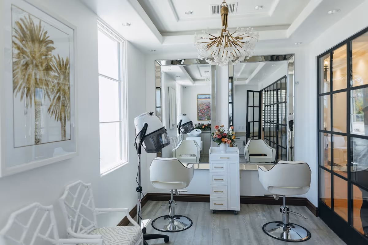 Bright salon-style room with two white styling chairs, a large mirror, a hooded hair dryer, a central drawer unit and a chandelier.