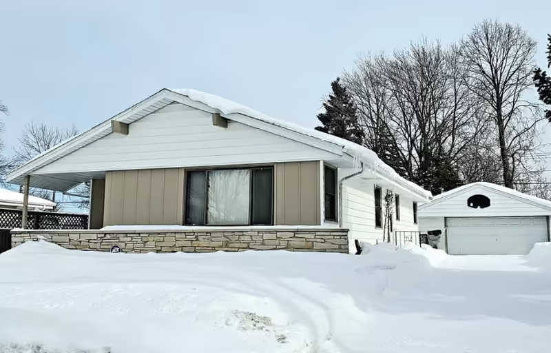 Snow-covered single-story house with stone accents and a detached garage.