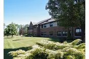 Exterior view of a two-story brick building with multiple windows, surrounded by green grass, trees, and shrubs under a clear sky.