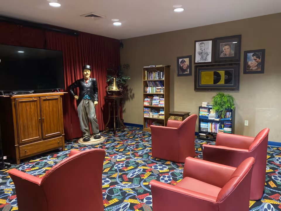 Seating area with four red armchairs facing a large TV, bookshelves, framed art and a full-body statue in a colorful carpeted room.