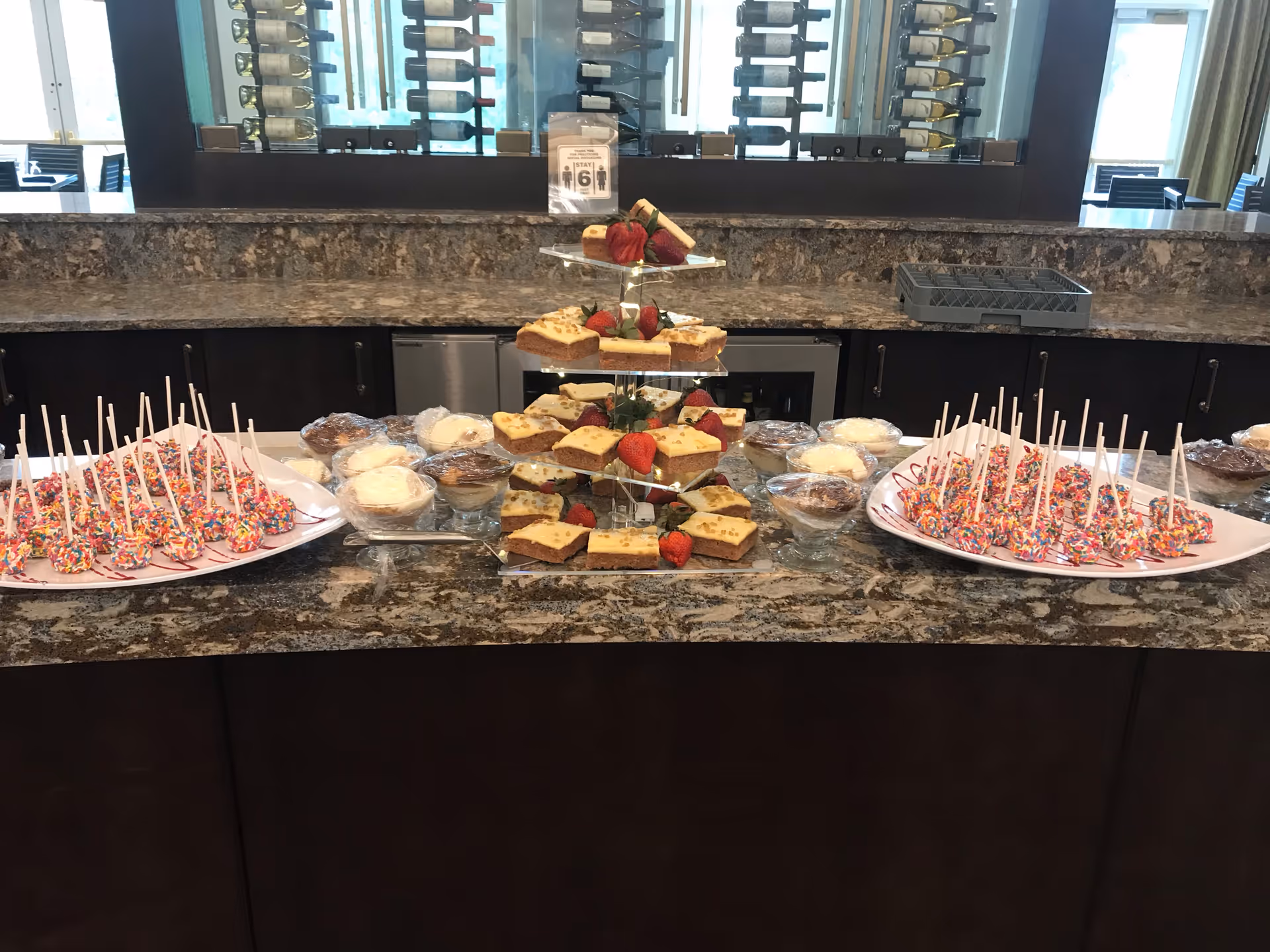 A dessert buffet setup on a granite countertop featuring plates of cake pops with colorful sprinkles, small glass bowls of various desserts, and a tiered stand with slices of cake garnished with strawberries. Behind the counter is a wine rack with bottles and a sign indicating a stay limit of 6 people.
