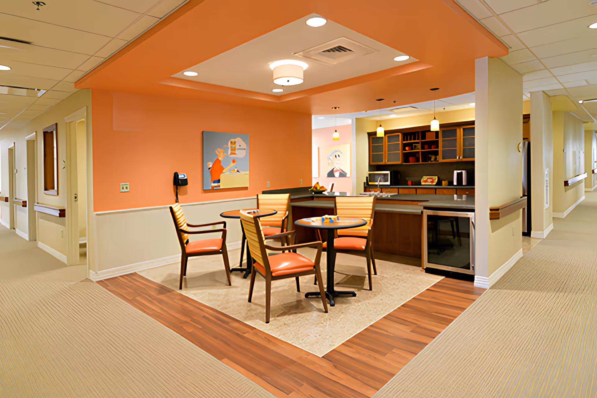 Open dining and kitchenette area with round tables, orange chairs, and a serving counter in a senior living facility.