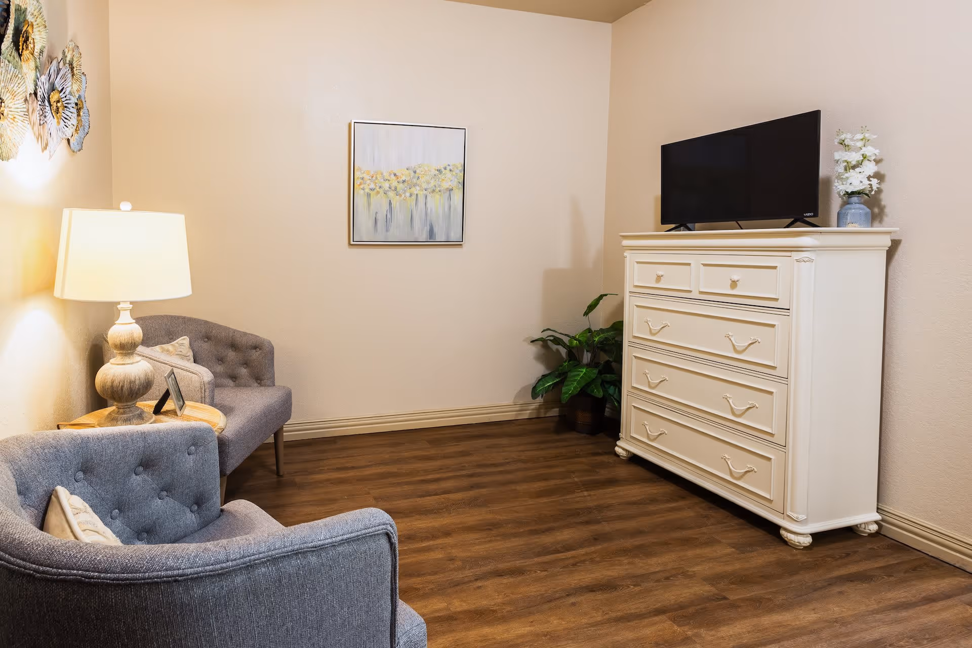 A cozy living room area with two gray upholstered armchairs, a wooden side table with a lamp, a white dresser with a flat-screen TV on top, a potted plant, and a framed painting on the wall. The room has wooden flooring and beige walls.