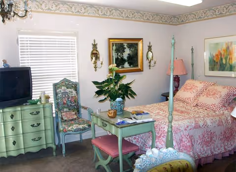 A cozy bedroom with a four-poster bed dressed in pink floral bedding. The room features a green dresser with a TV on top, a matching green desk with a pink cushioned bench, and a decorative chair with floral upholstery. There are framed paintings and wall sconces on the walls, a window with white blinds, and a pink lamp on a bedside table.