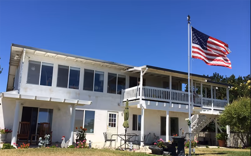 Two-story white senior living facility with a balcony, front patio, and American flag.