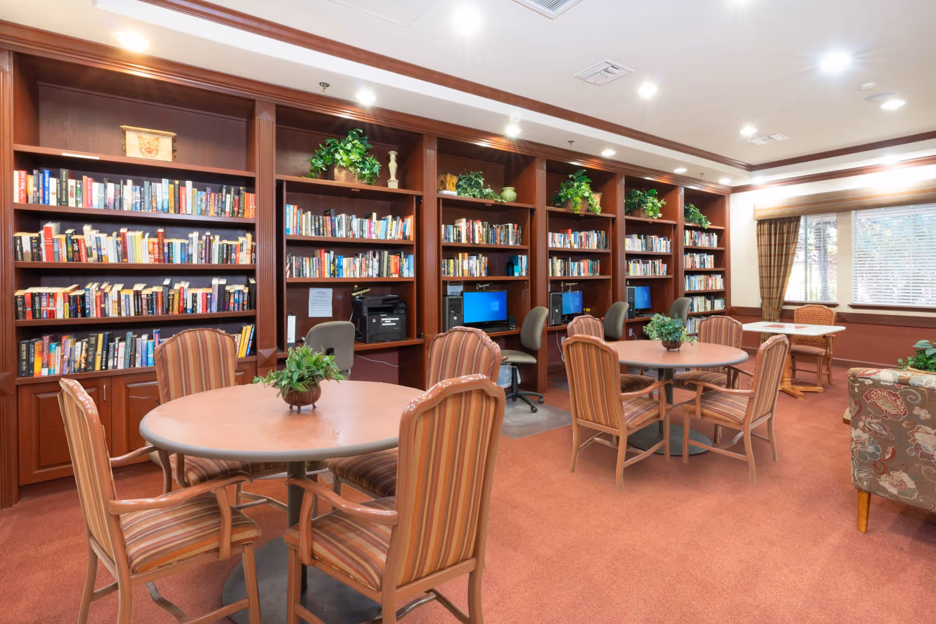 A cozy library and computer room with wooden bookshelves filled with books and decorative plants. There are several round tables with striped upholstered chairs around them, and a few computer workstations with chairs along the bookshelves. The room has warm lighting, carpeted floors, and a window with blinds and curtains letting in natural light.