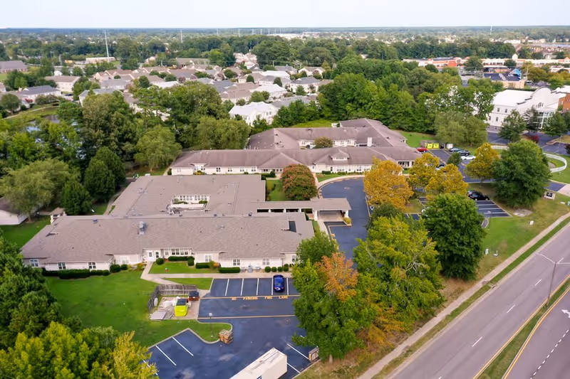 Aerial view of Chesapeake Place Senior Living facility showing multiple connected buildings with gray roofs surrounded by green trees and lawns. There is a parking lot with several cars and a road running alongside the property. Residential houses and more greenery are visible in the background.