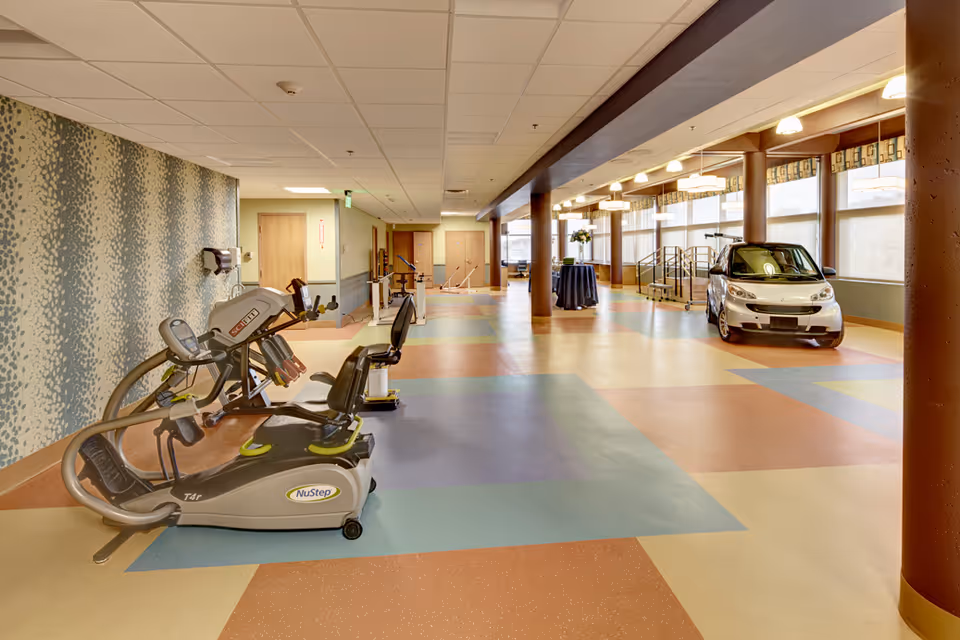A spacious rehabilitation room with exercise equipment including a NuStep machine on the left and a small silver car on the right. The room has a colorful patterned floor, large windows with curtains, and several ceiling lights. There are also handrails and a small stage area in the background.