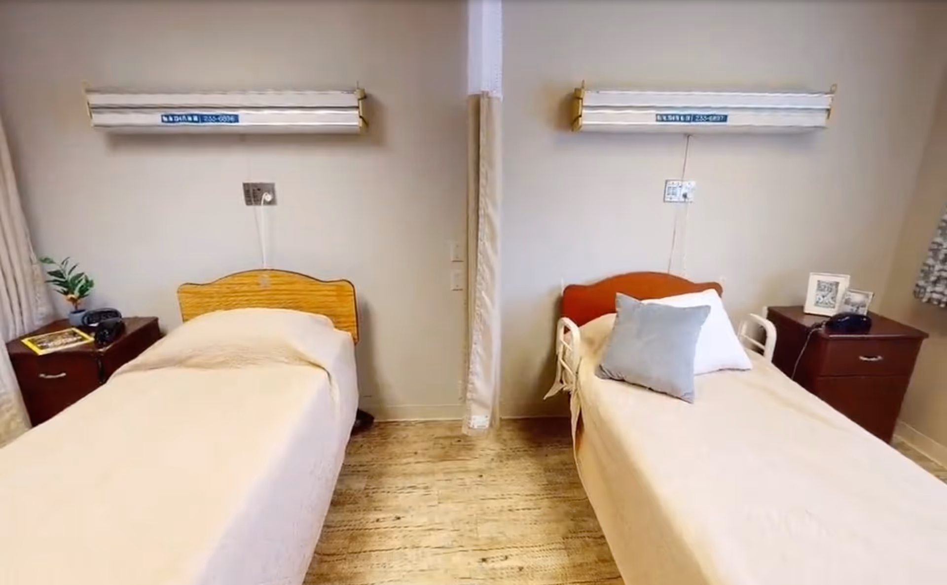 Two single beds with bedside tables and overhead medical light rails separated by a center privacy curtain in a shared senior bedroom.