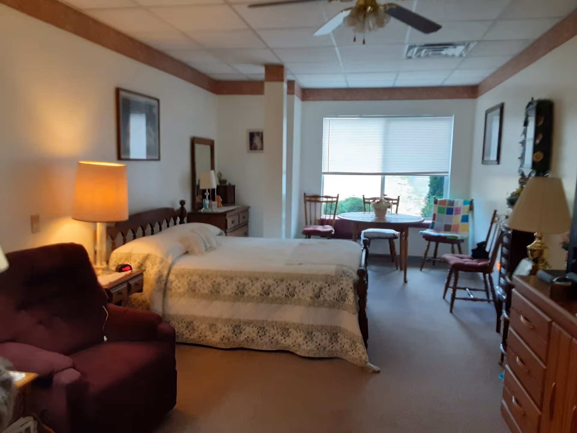 A cozy bedroom in a senior living facility with a bed covered in a patterned quilt, a wooden headboard, and a bedside table with a lamp and alarm clock. There is a burgundy armchair to the left, a dresser with a mirror, and a small round table with chairs near a window with blinds. The room has beige walls, carpeted floor, and ceiling tiles with a ceiling fan.