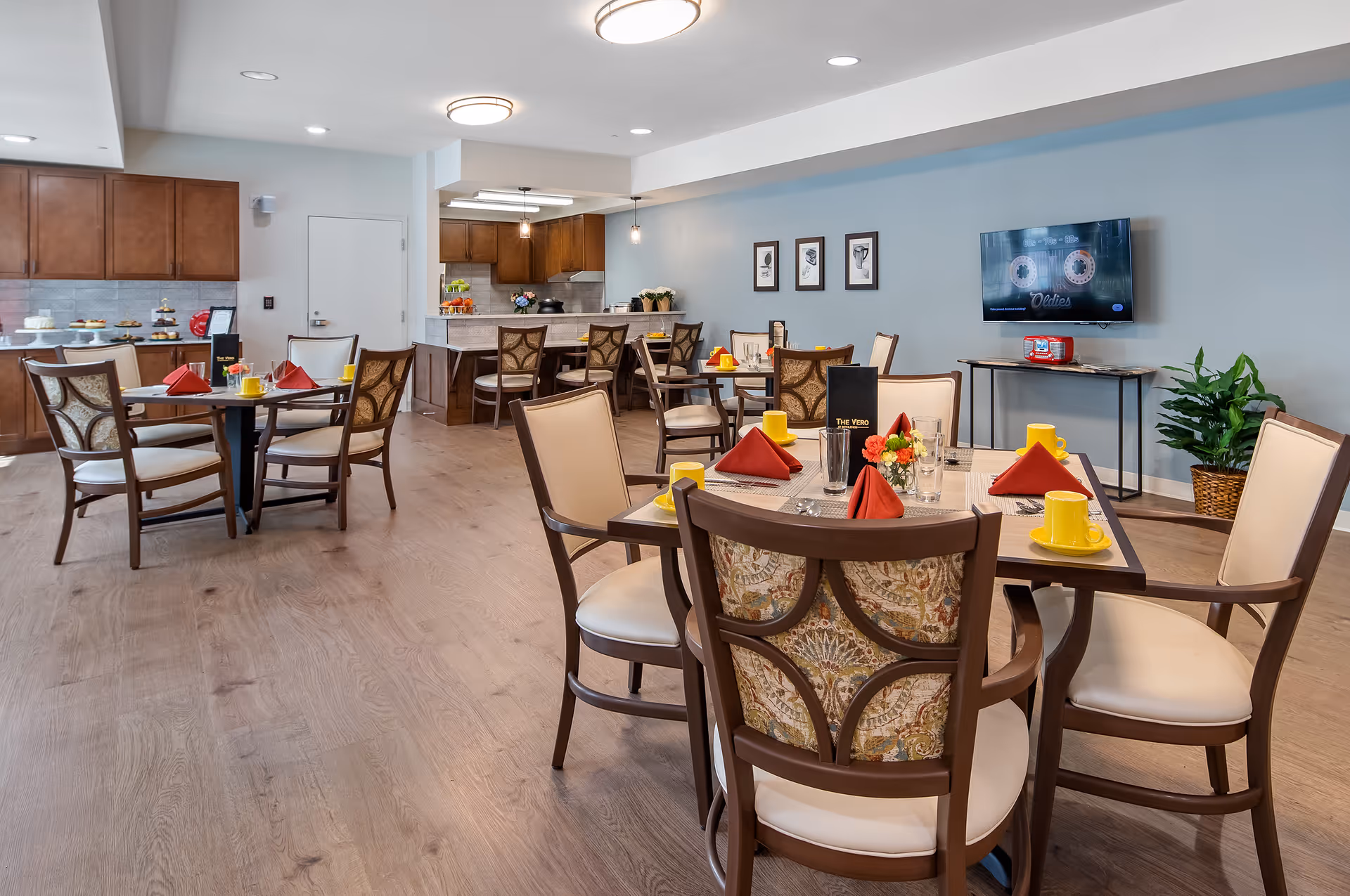 A bright and spacious dining area in a senior living facility with several tables set with yellow cups, red napkins, and floral centerpieces. The room features wooden chairs with patterned backs, light wood flooring, and a light blue wall with framed artwork. In the background, there is a kitchen area with wooden cabinets and a counter with bar stools. A flat-screen TV is mounted on the wall above a small table with a red radio and some magazines. A green plant in a woven basket is placed near the TV.