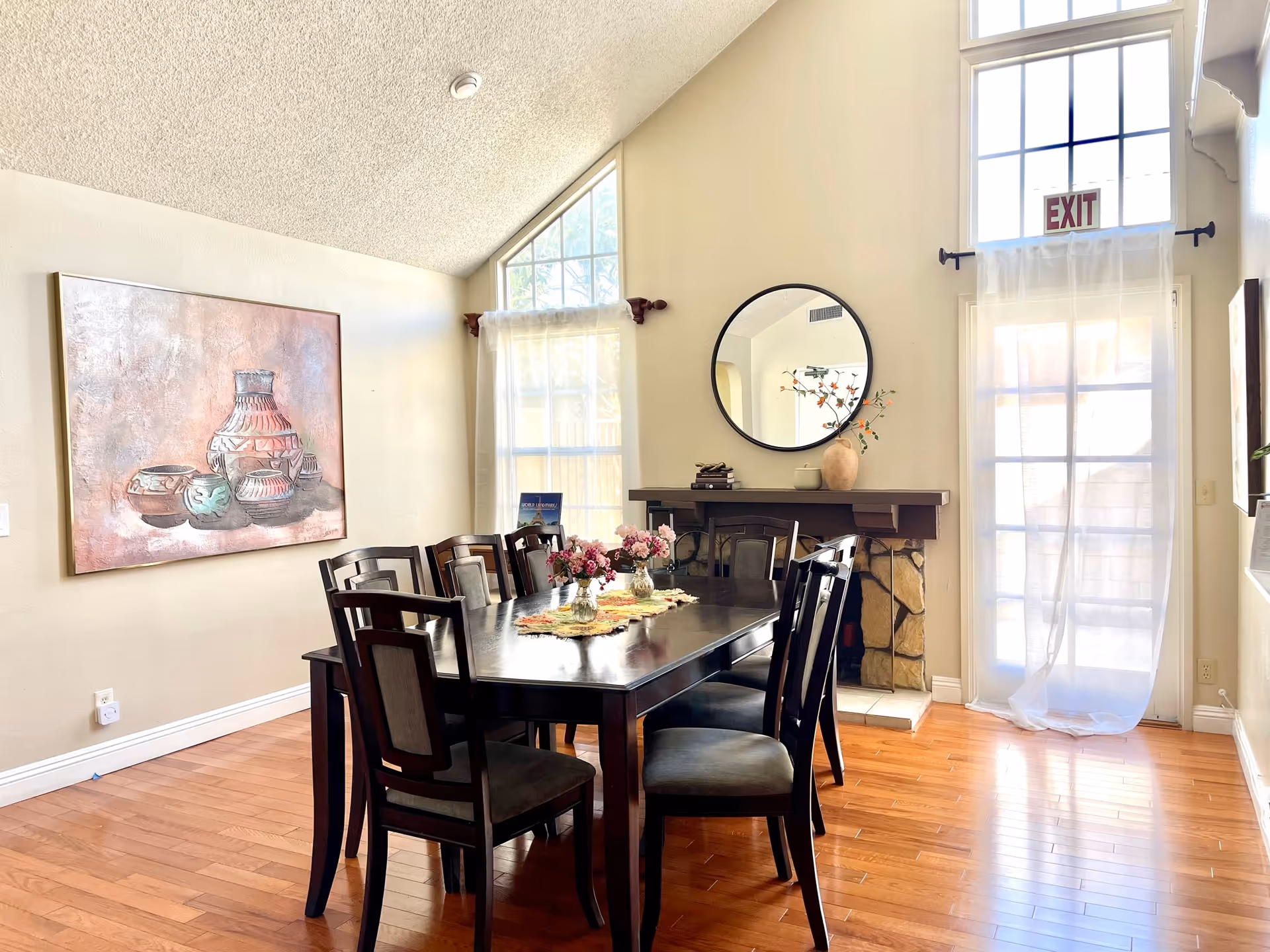 Sunlit dining room with a long dark wood table and chairs, a round mirror above a fireplace, wall art, and tall windows with sheer curtains.