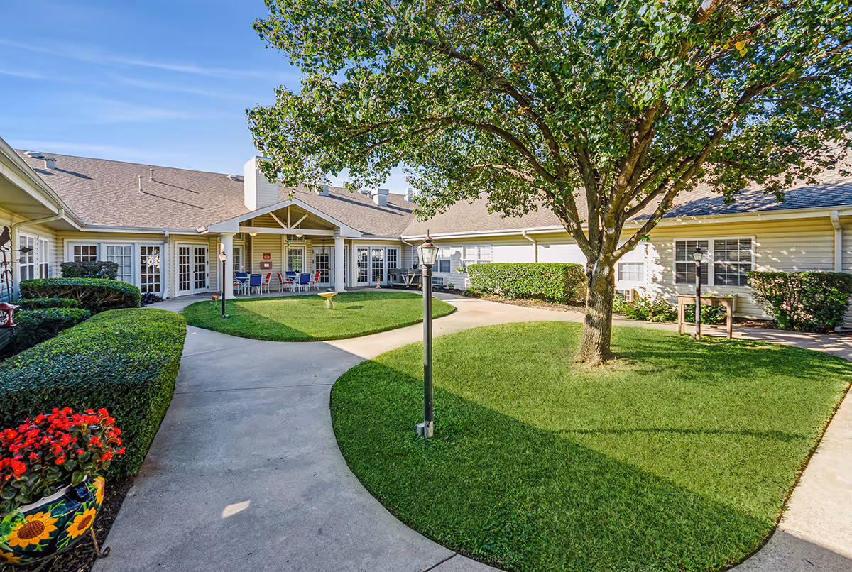 Well-maintained courtyard with a central tree, paved walkways, green lawn and surrounding single-story senior living units with a covered seating area.